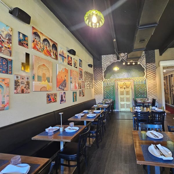 Restaurant interior with a long banquette, wooden tables, and colorful Indian-inspired artwork on the walls.