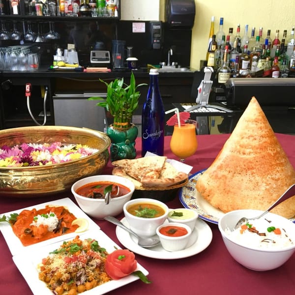 A table laden with various Indian dishes, including dosa, curries, rice, naan, and a drink.