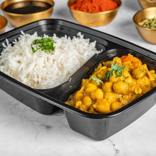 Indian chickpea curry and white rice in a black takeout container, with spices in golden bowls.